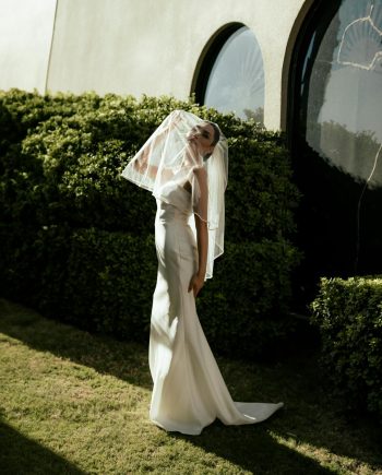 A beautiful bride in a white gown poses gracefully outdoors beneath sunlight.